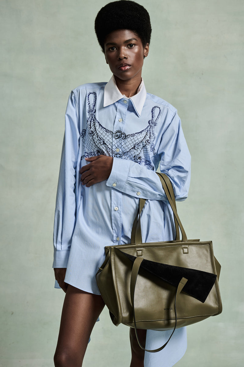 MONSE Spring 2026 Collection look 16 person wearing a light blue shirt with a graphic design, holding a green handbag against a neutral background