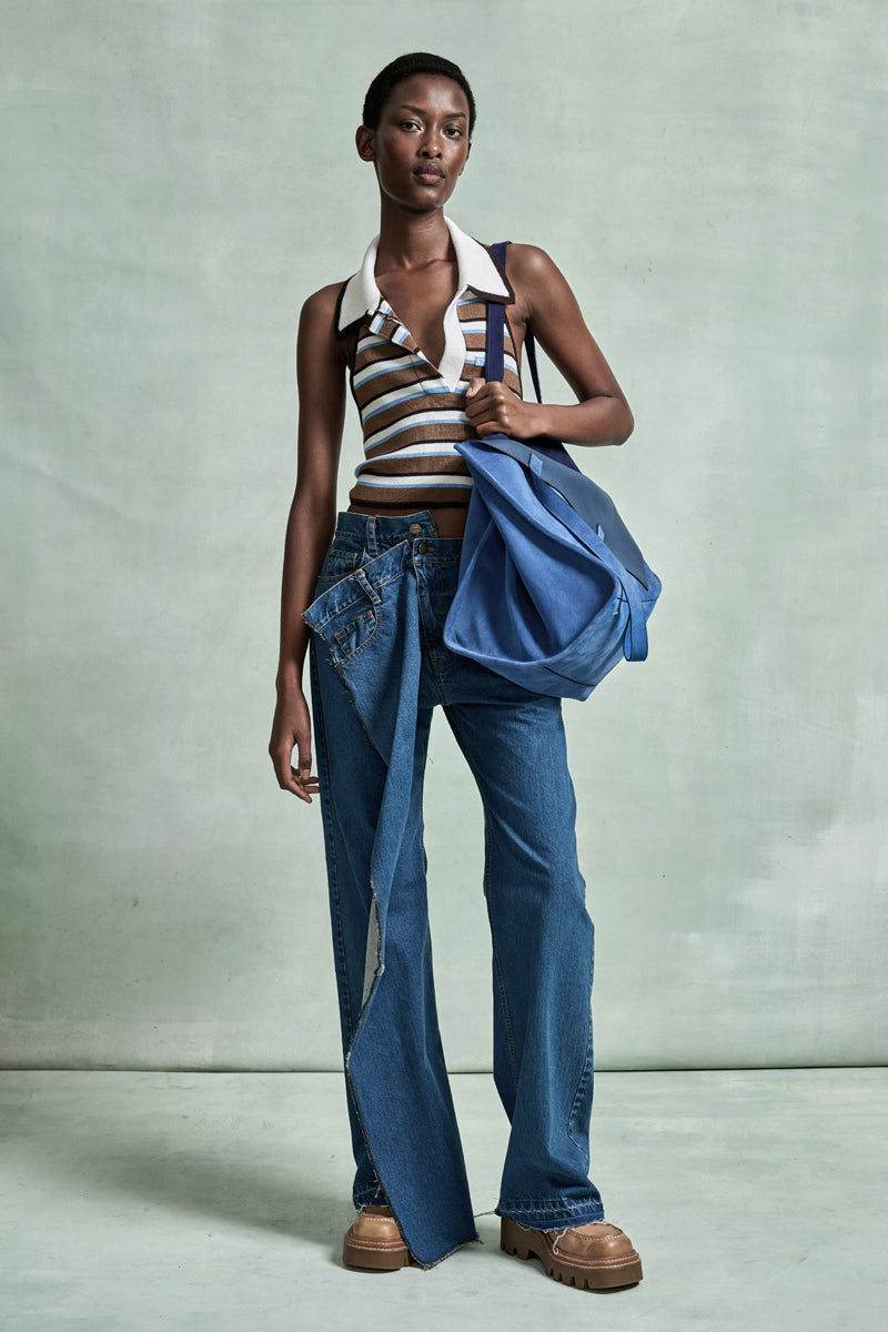MONSE Spring 2026 Collection look 20 woman wearing a striped top and blue jeans, holding a blue bag against a light gray background