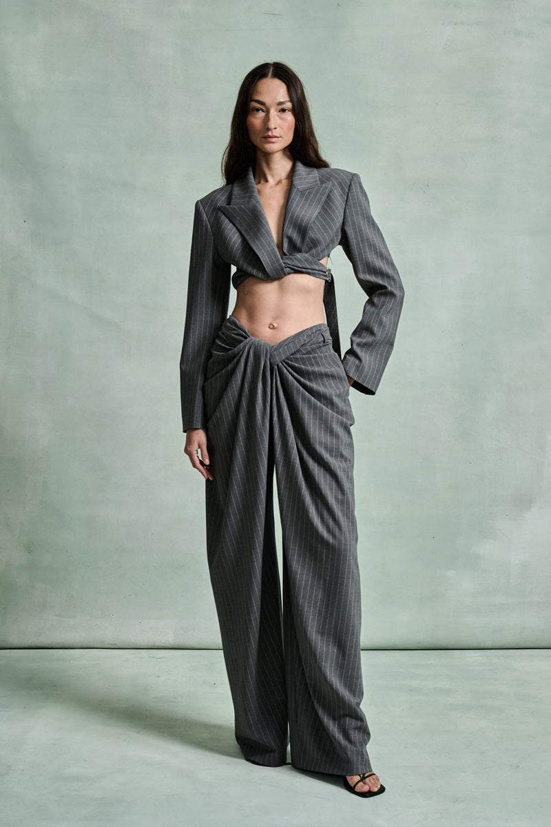 MONSE Spring 2026 Collection look 6 woman wearing a gray pinstripe suit with a cropped blazer and wide-leg pants on a light green background