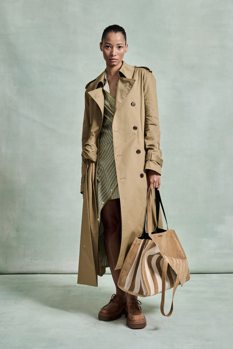 MONSE Spring 2026 Collection look 9 woman wearing a beige trench coat holding a patterned handbag against a light green background